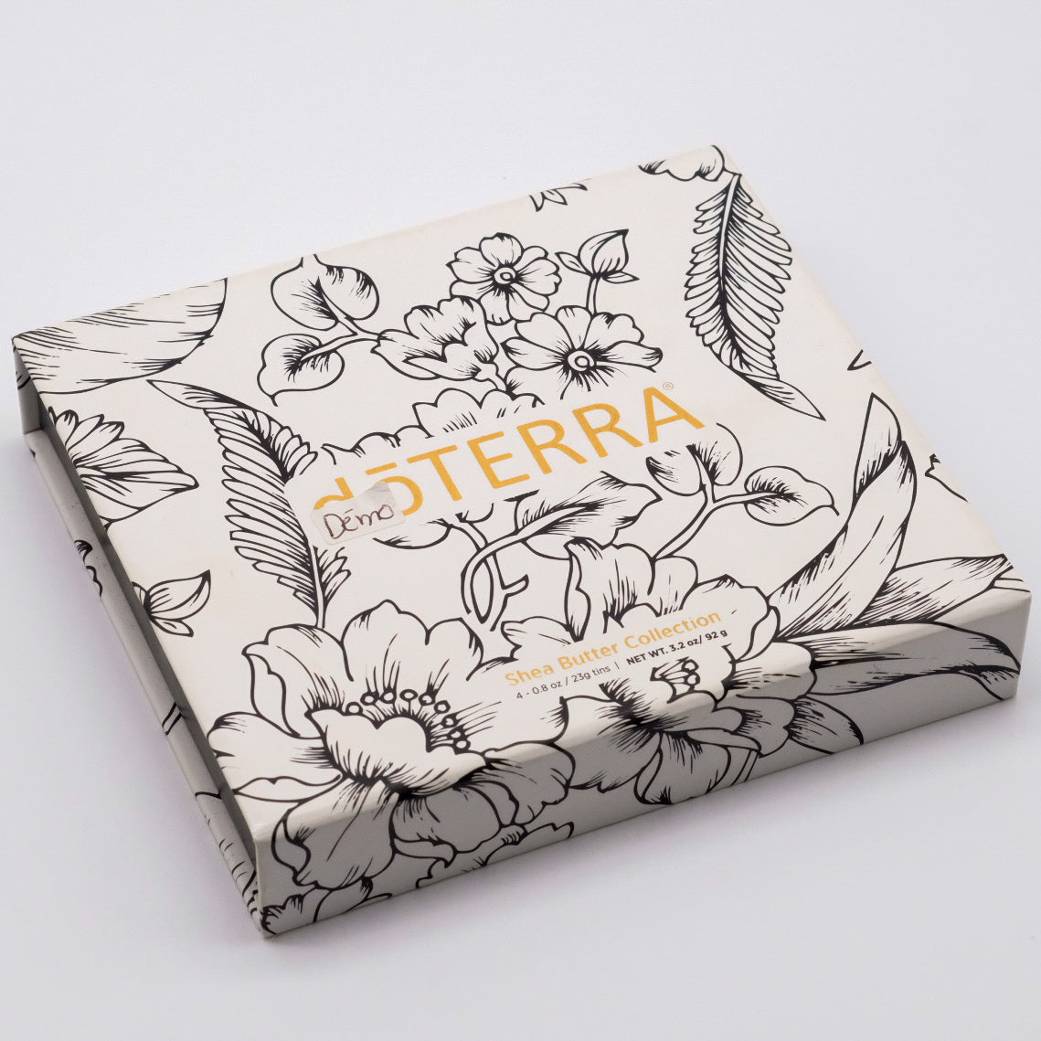 Coffret fermé blanc doTERRA Collection beurre de karité, avec logo doré et motif floral noir.