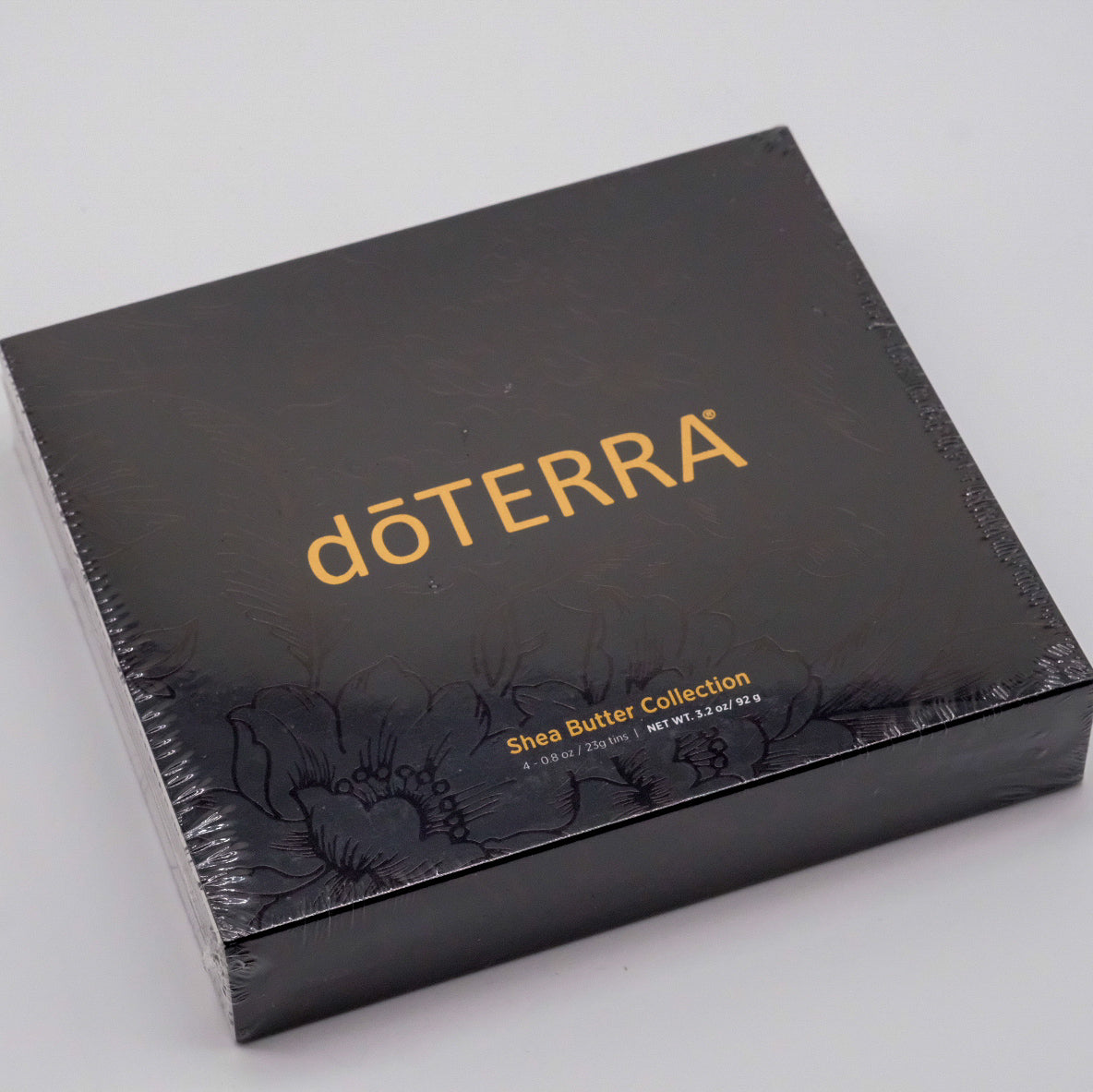 Coffret noir doTERRA Collection beurre de karité sous emballage plastique, avec logo doré et motif floral ton sur ton.