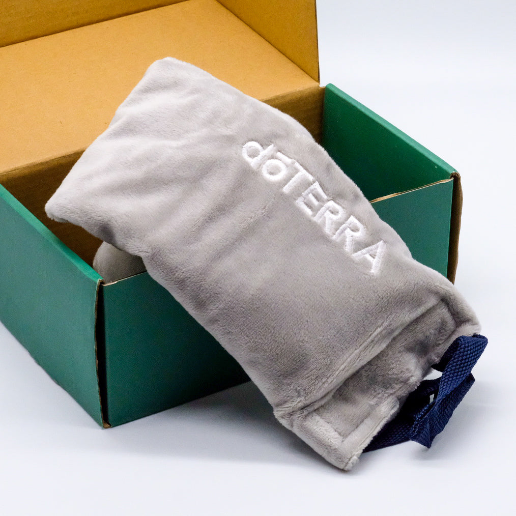 Coussin thermique gris doTERRA pour le cou et le dos, plié dans une boîte ouverte avec sangle bleu foncé visible.
